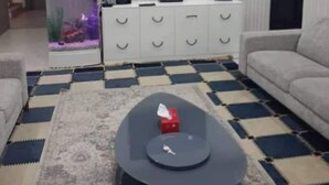 Een moderne woonkamer met een flatscreen televisie aan de muur, een witte entertainmentkast, een kamerkleed met een patroon en een ronde salontafel met een afstandsbediening.
