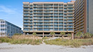 Ein mehrstöckiges Hotel mit Balkonen, einem überdachten Parkbereich und einem Sandstrand im Vordergrund.