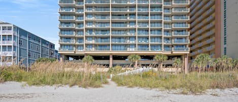 A multi-story hotel with balconies, a covered parking area, and a sandy beach in the foreground.