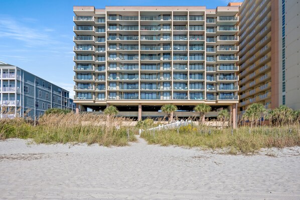 A multi-story hotel with balconies, a covered parking area, and a sandy beach in the foreground.