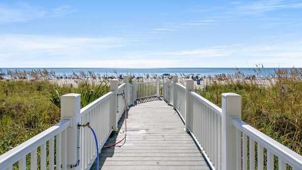 Ein hölzerner Steg führt zu einem Strand bei strahlendem Himmel.