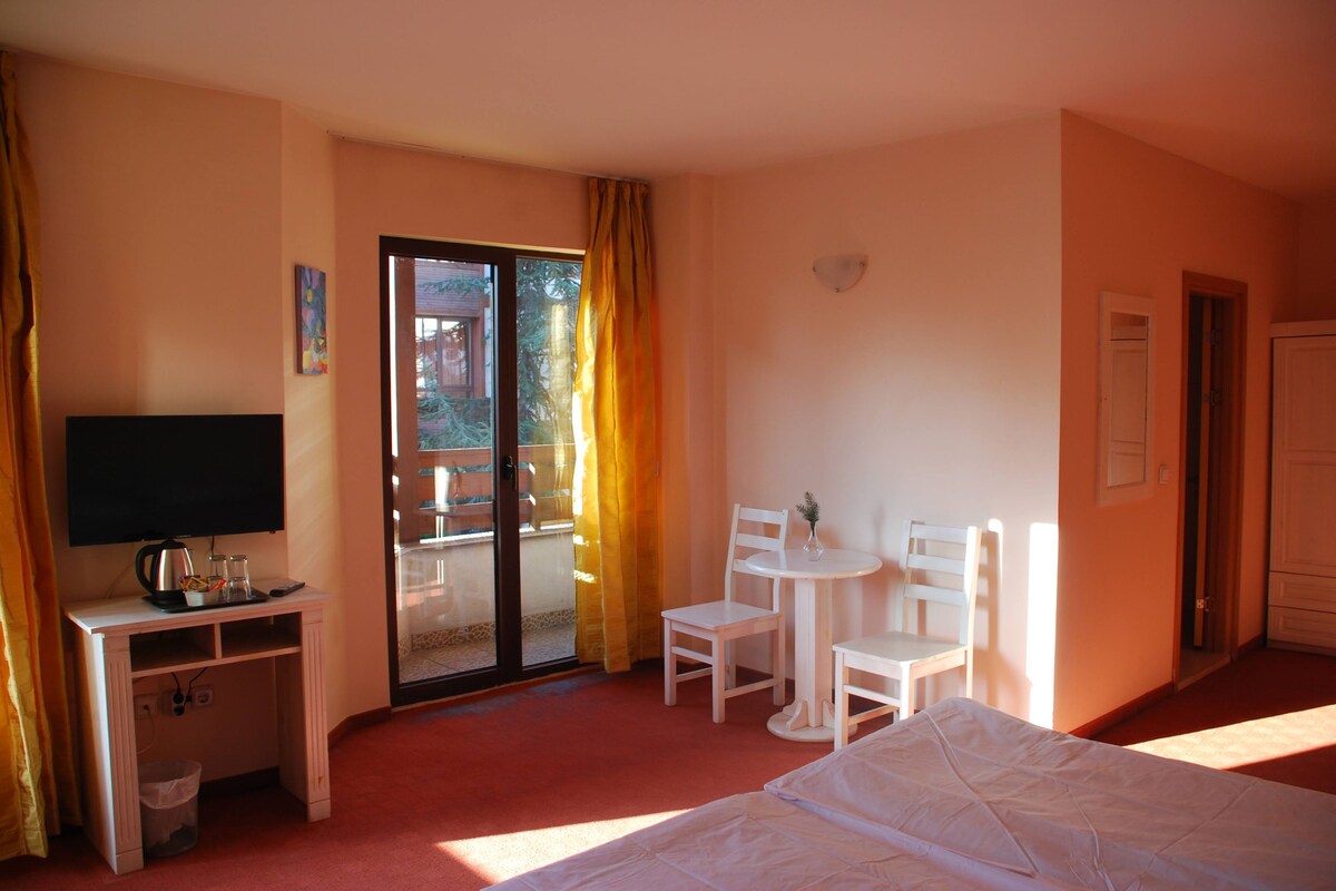 A hotel room with a bed, a desk with a television, a small table with chairs, and a sliding glass door to the balcony.