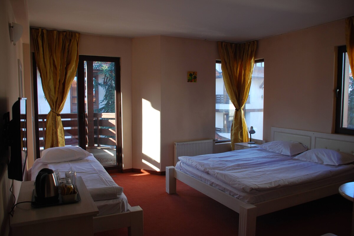 A hotel room with two beds, a desk with a kettle, a radiator, and a window with curtains.