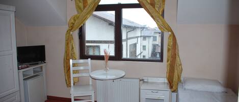 Una habitación pequeña y bien iluminada con una cama individual, un escritorio con silla, un televisor y una ventana con cortinas.