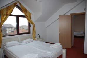 A bedroom with a bed, a nightstand, a lamp, a window with curtains, and a door leading to another room.