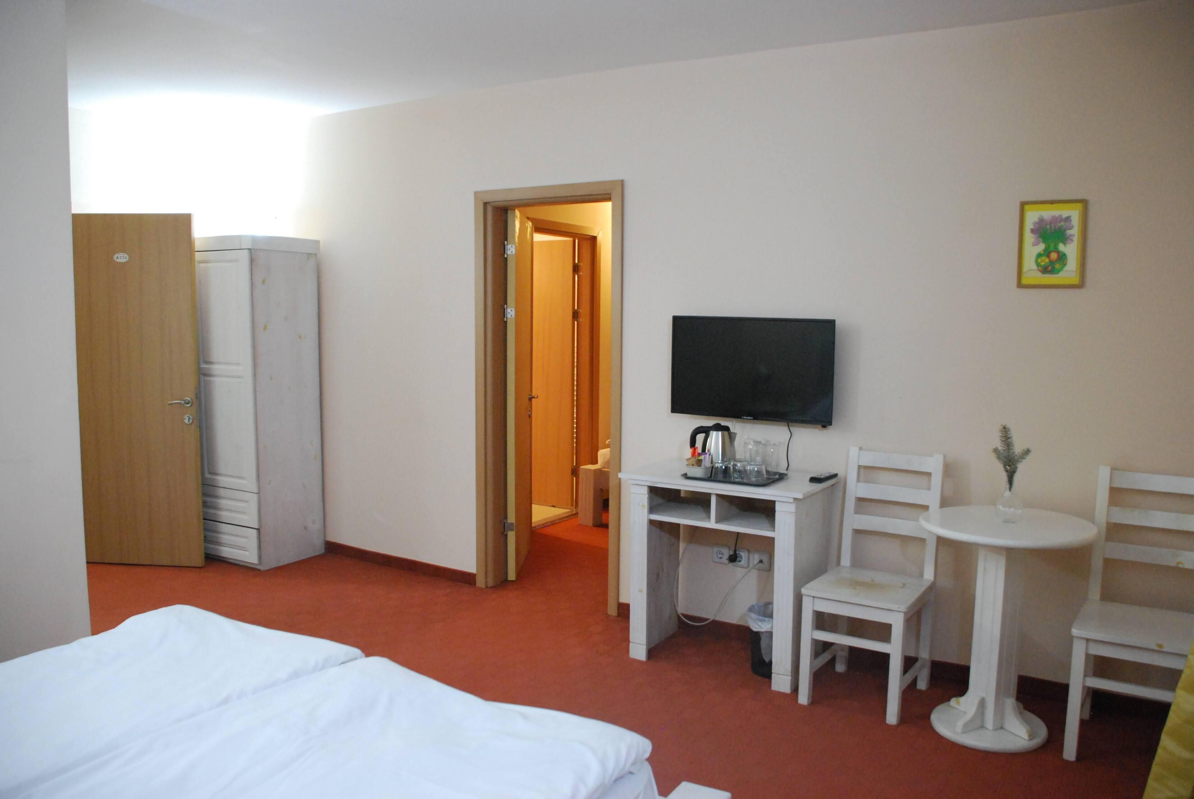 A hotel room with a bed, a desk with a TV, two chairs, a small table, and a door leading to another room.