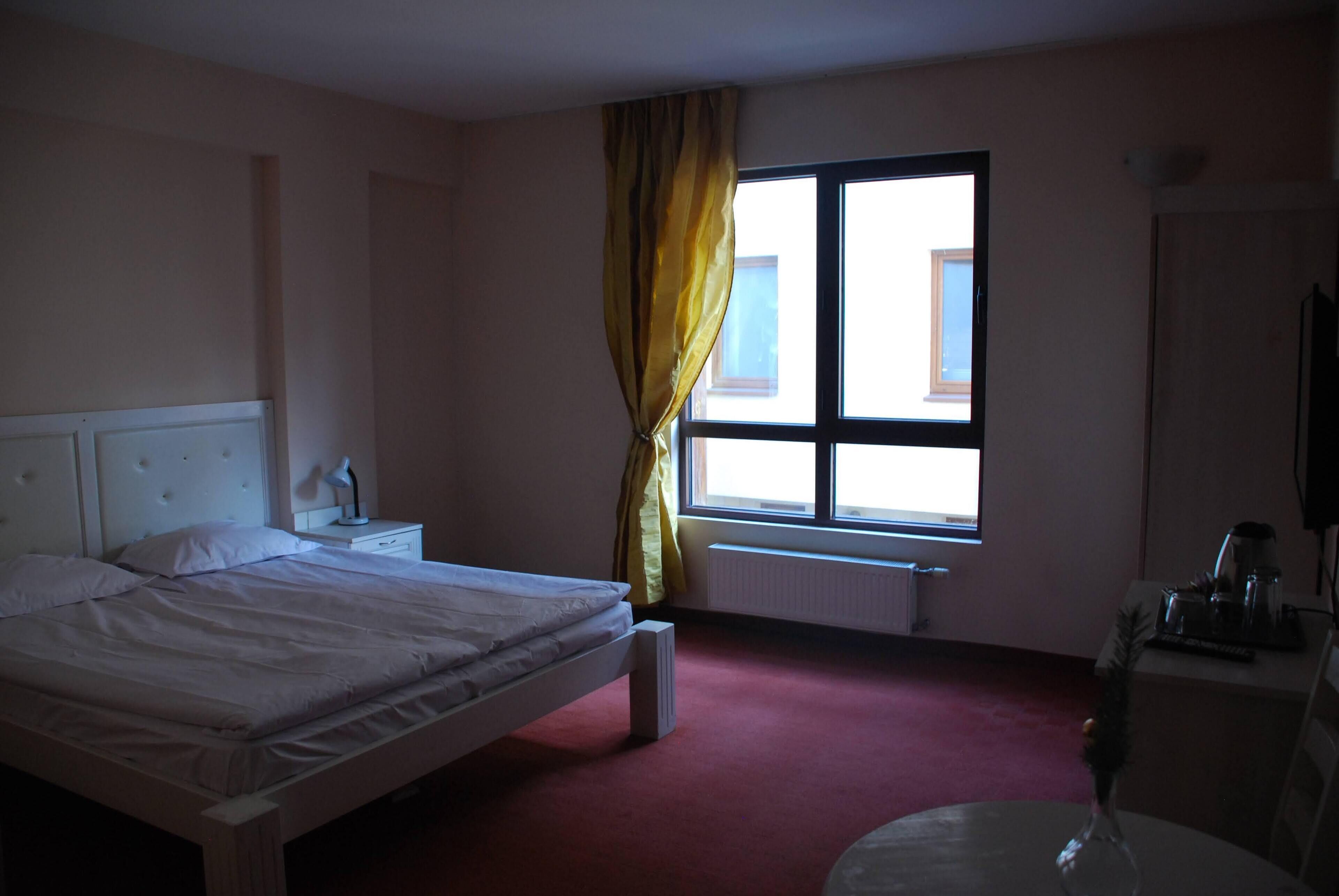 Quarto de hotel com cama, mesa de cabeceira, janela com cortinas, radiador e uma cadeira.