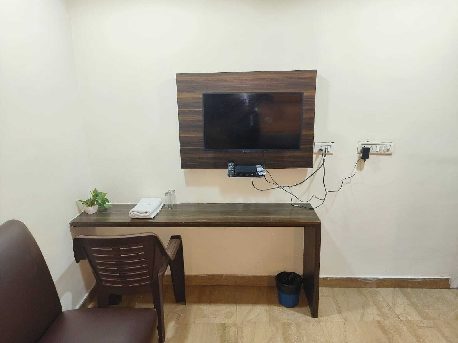 A small, neatly arranged room with a wooden desk, a chair, a television mounted on the wall, and a trash bin.