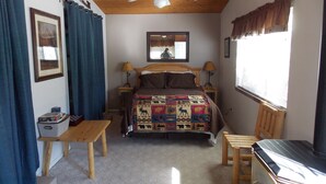 A bedroom with a wooden ceiling, a bed with a patterned blanket, two bedside lamps, a wooden nightstand, a small table, a window with curtains, and a ceiling fan.