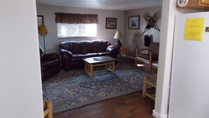 A living room with a brown leather sofa, a wooden coffee table, and a patterned rug. There is a window with blinds, a lamp, and a framed picture on the wall.