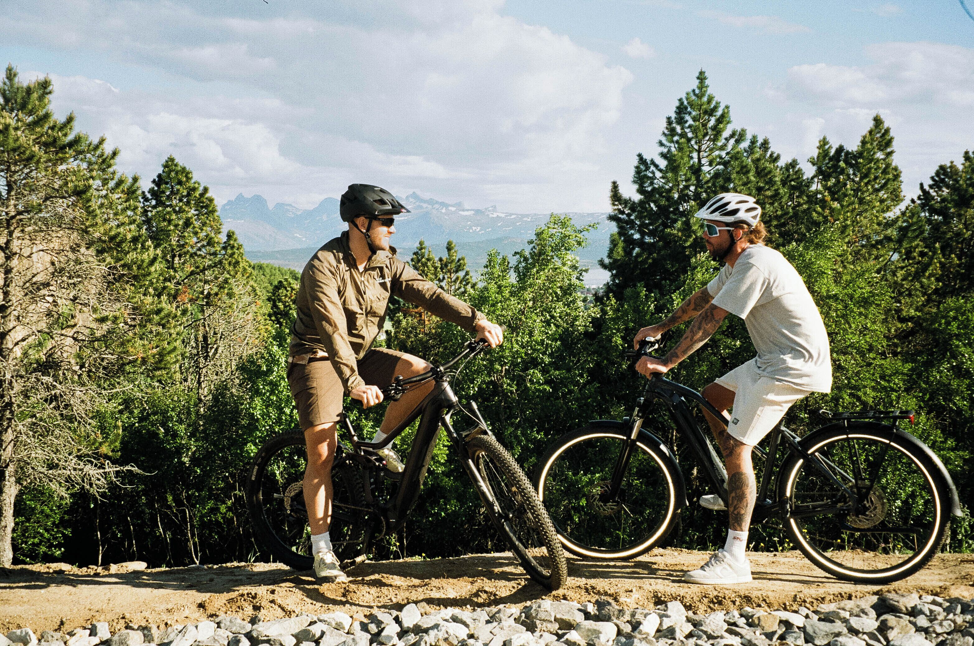 Two people on bicycles in a natural setting with trees and mountains in the background.
