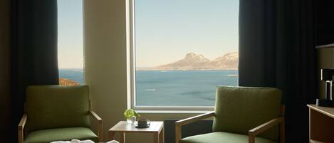 A hotel room with two green chairs, a bed, a small table with a cup and a flower, and a view of the sea and mountains through a large window.