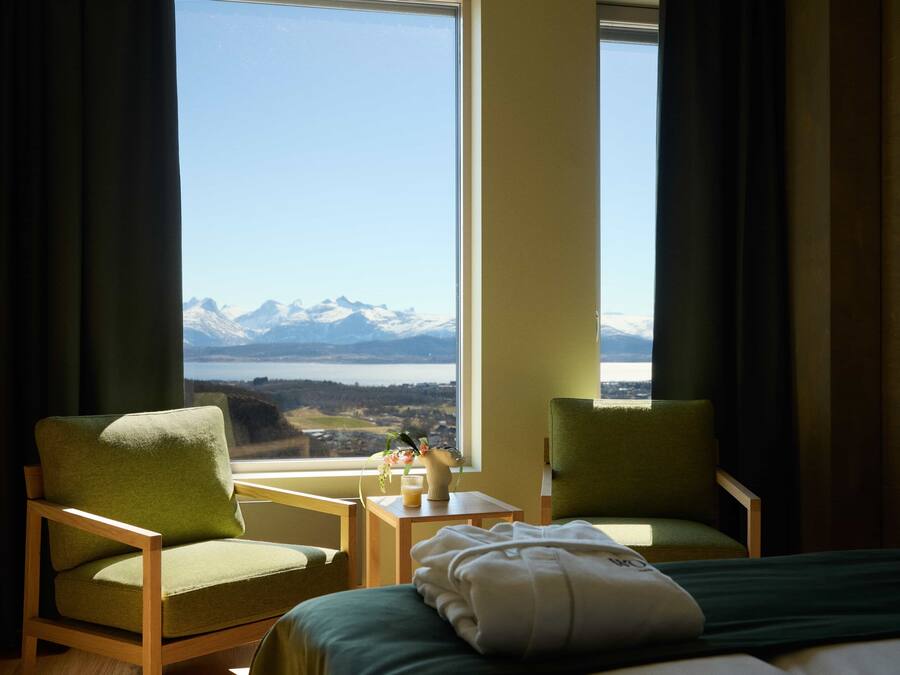 A hotel room with two green armchairs, a bed with a green bedspread, and a view of mountains and a lake through large windows.