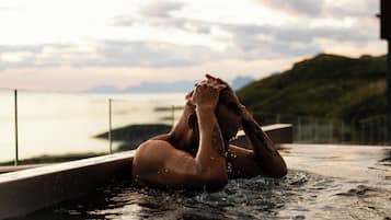 Una persona en un jacuzzi con vista a un cuerpo de agua y colinas al fondo.