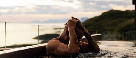 A person in a hot tub with a view of a body of water and hills in the background.