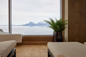 Habitación con un ventanal grande que ofrece vistas a montañas y agua. Hay una planta en maceta sobre una mesa de madera.