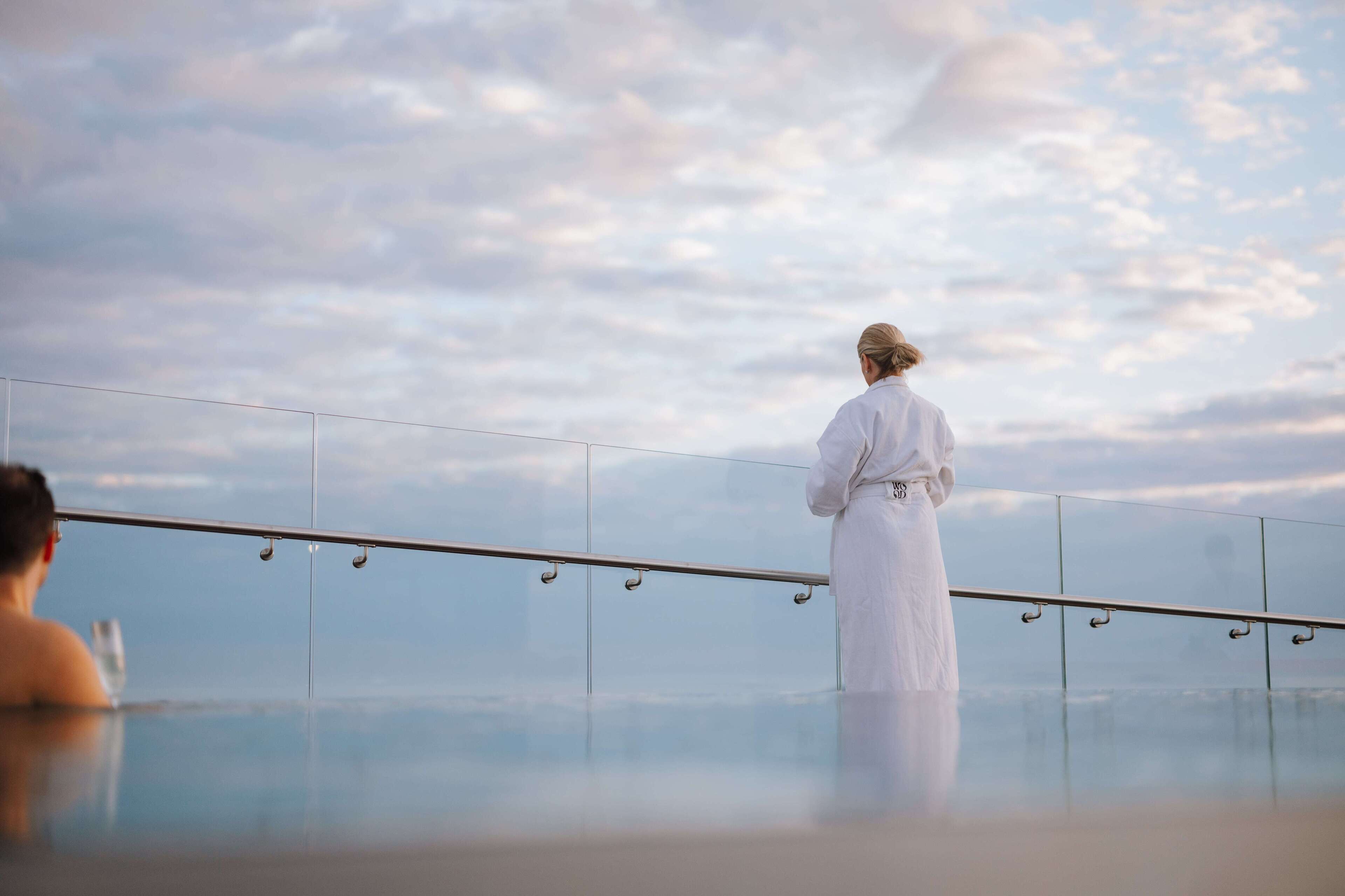 A person in a white robe standing by a glass railing overlooking a body of water with a cloudy sky in the background.