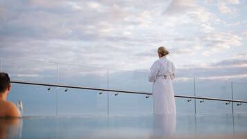 A person in a white robe standing by a glass railing overlooking a body of water with a cloudy sky in the background.