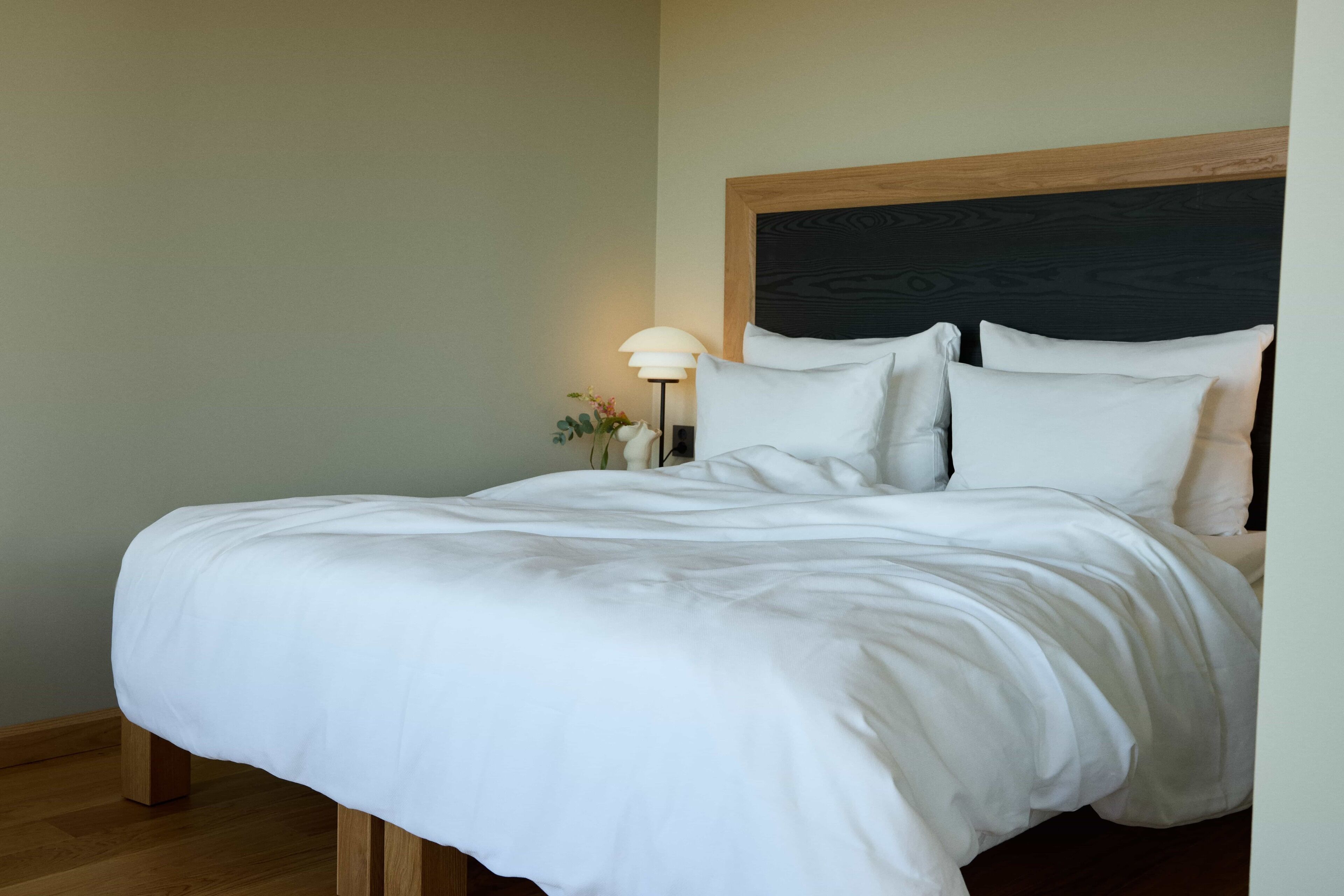 Cama bem arrumada com roupa de cama branca, cabeceira de madeira e abajur com cúpula branca.