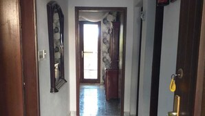 Un couloir avec un sol carrelé, une porte en bois sur la gauche, un miroir au mur et un extincteur sur la droite.
