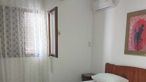 Une chambre avec un lit en bois, du linge de lit blanc, une table de chevet, une lampe, une fenêtre avec des rideaux légers et un appareil de climatisation.