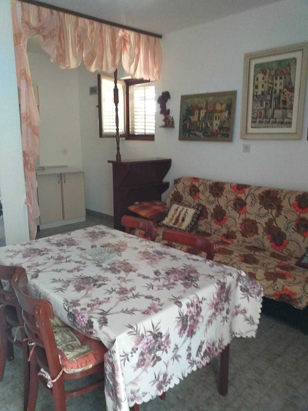 A living room with a floral tablecloth-covered dining table, a sofa with a floral pattern, and a window with blinds.