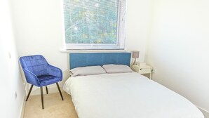 Ein kleines, sauberes Zimmer mit einem Einzelbett, einem blauen Stuhl, einem Nachttisch mit Lampe und einem Fenster mit Jalousien.