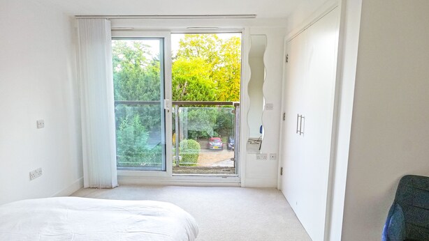 Ein Schlafzimmer mit Bett, einer Schiebetür zum Balkon und Blick auf Bäume sowie ein Auto vor dem Haus.