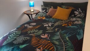 Een slaapkamer met een bed dat is voorzien van een bedsprei met een tropisch bladmotief, een rond nachtkastje met een blauwe lamp en een enkel geel kussen op het bed.