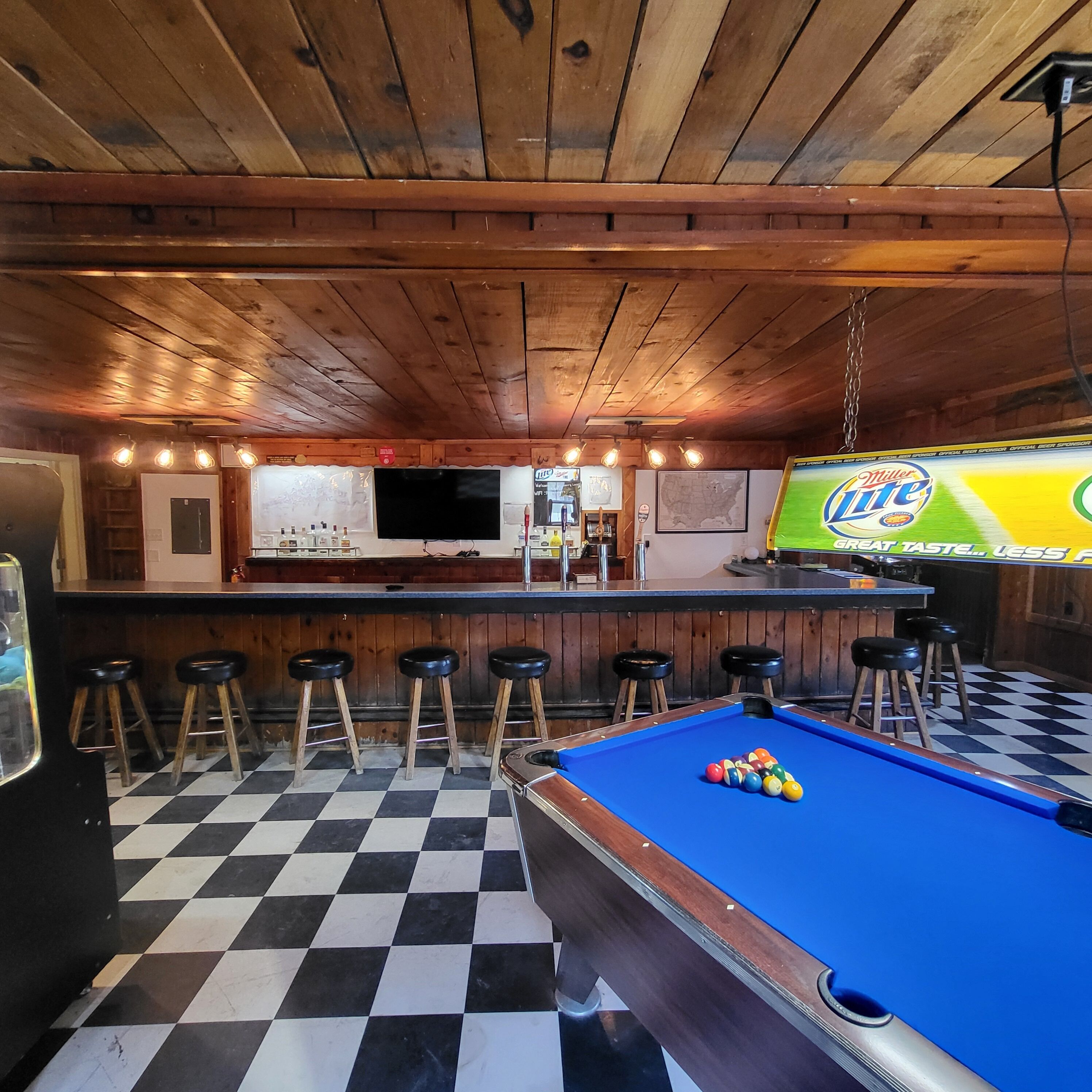 Una mesa de billar con bolas, una zona de bar con taburetes y una pantalla de televisión mostrando un anuncio.