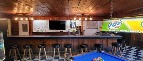 Una mesa de billar con bolas, una zona de bar con taburetes y una pantalla de televisión mostrando un anuncio.