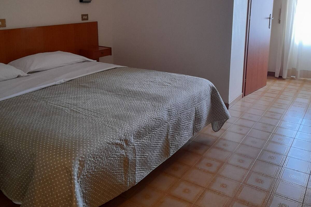 A hotel room with a bed, a nightstand, a wall-mounted cross, and a door with curtains.