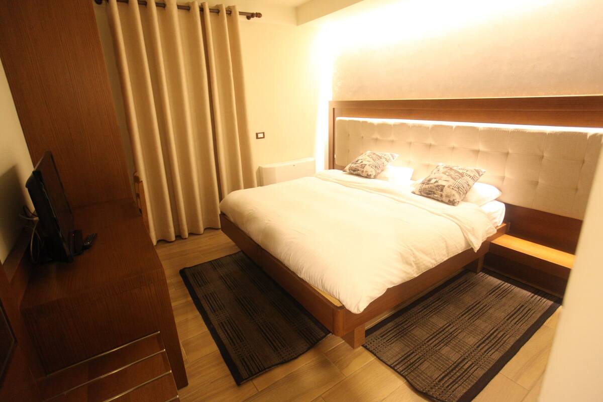 A hotel room with a bed, a wooden nightstand, a TV, and a window with curtains.