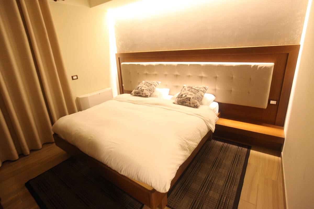 A neatly made bed with a wooden headboard and two pillows, a dark rug, and a light-colored wall.