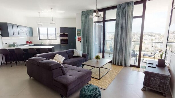 Un salon moderne avec un canapé, une table basse et un pouf. On aperçoit une cuisine à l’arrière-plan, avec des tabourets de bar.