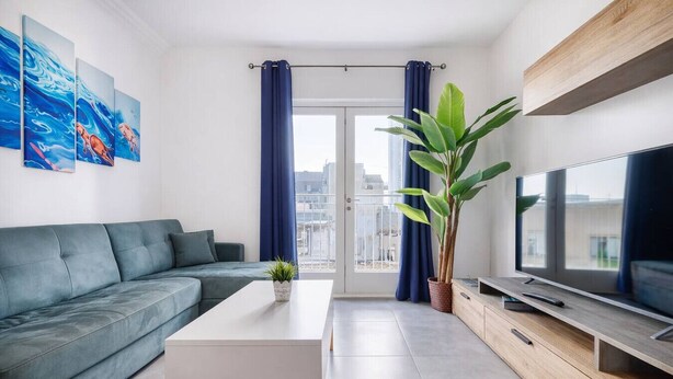 A modern living room with a grey sofa, a white coffee table, a large potted plant, a TV, and a balcony with blue curtains. - Modern 3-bedroom apartment Pieta (Tal-Pietà)