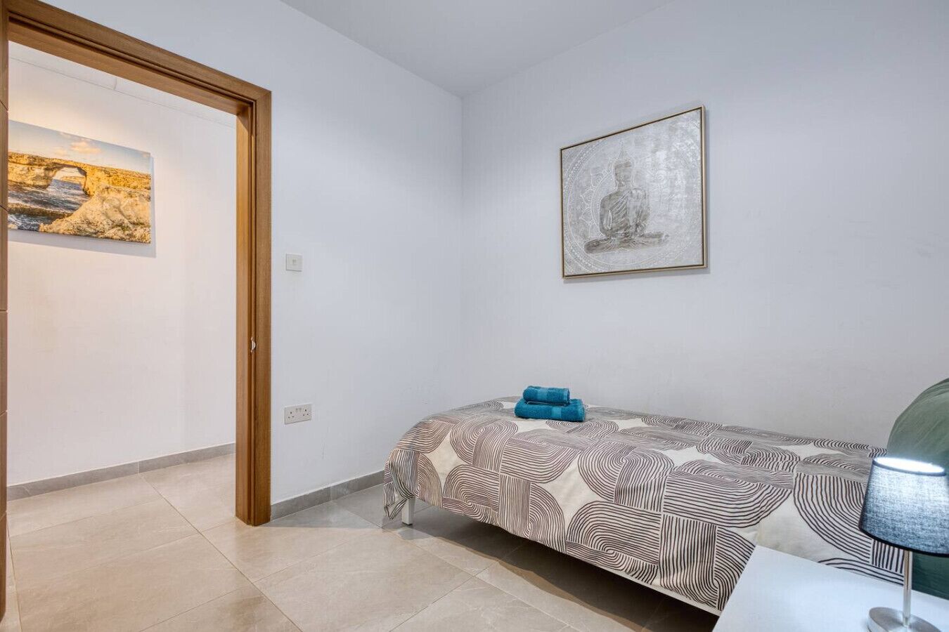 A bedroom with a bed, a wall-mounted lamp, a framed artwork, and a door leading to another room.