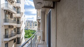Un balcón estrecho con barandilla de metal, vistas a edificios cercanos y una puerta a la derecha.