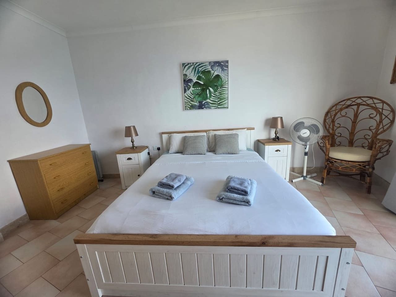 Ein Schlafzimmer mit einem Bett, zwei Nachttischen, einem Wandventilator, einer Holzkommode, einem Korbstuhl und einem Gemälde.