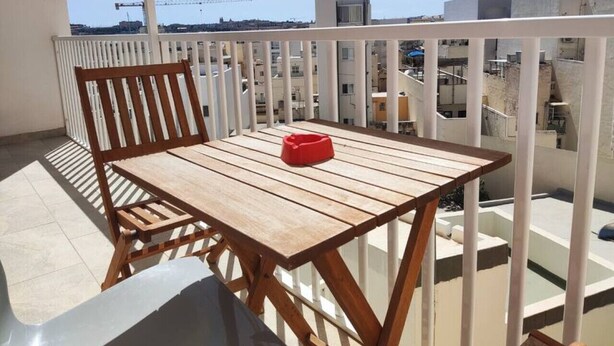 A balcony with a wooden table and chair, a red object on the table, and a view of buildings and construction cranes in the background. - Modern 2-bedroom apartment in Gzira (Il-Gżira)