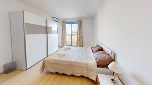 A modern bedroom with a large bed, a wardrobe, and a view of the city. - Modern 5th floor 2 bedroom apartment in Tigne (Sliema)