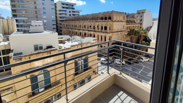 Ein Balkon mit Blick auf eine Stadtlandschaft, darunter moderne und historische Gebäude, ein Parkplatz und ein klarer Himmel.