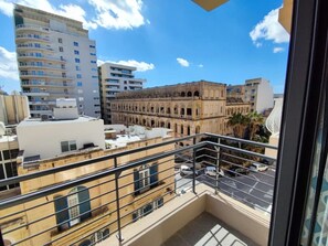 Ein Balkon mit Blick auf eine Stadtlandschaft, darunter moderne und historische Gebäude, ein Parkplatz und ein klarer Himmel.