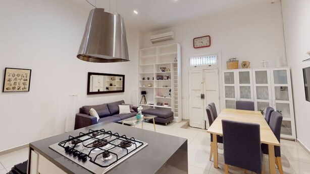 A modern kitchen with a gas stove, a dining area with a table and chairs, a sofa, and a bookshelf. - Spacious 3 bedroom maisonette (Tas-Sliema)