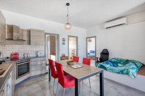 A modern kitchen with a dining area, a bed with a blue and green patterned blanket, and a black gaming chair.