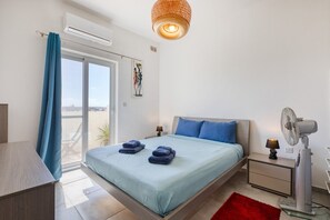 A bedroom with a bed, a desk, a chair, a window with curtains, a ceiling light, a bedside table with a lamp, and a floor lamp. - the Queen Freddy Penthouse (Ta` Xbiex)