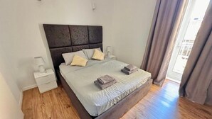 Un dormitorio con suelo de madera, una cama con cabecera, dos mesitas de noche y un ventanal con cortinas.