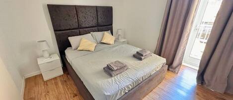 A bedroom with a wooden floor, a bed with a headboard, two bedside tables, and a window with curtains.