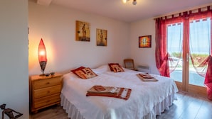A bedroom with a bed, a nightstand, a lamp, artwork on the wall, and a view through the window. - Chalet Panoramique Avec Sauna Avec vue sur le LAC Leman et Montagnes (Évian-les-Bains)