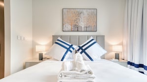 Un lit bien fait, avec du linge de lit blanc et des coussins bleus, une serviette pliée posée sur le lit et une œuvre d’art encadrée au-dessus du lit.